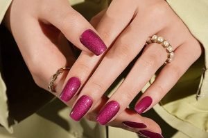 Glossy magenta gel nail strips on hands with gold and pearl rings, styled to match an elegant outfit